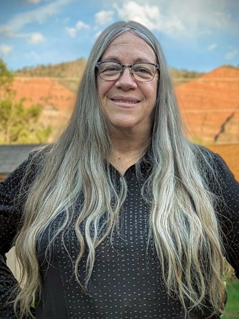 A mature woman with long hair standing in front of red hills in the background.